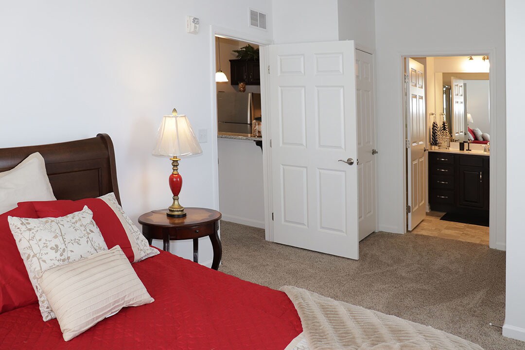 Bedroom with a red quilt and bedside lamp, looking through to the kitchen and bath